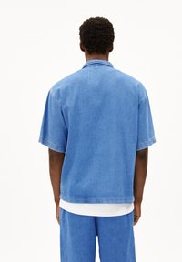 Leren spijkerhemd in lichtblauw, korte mouwen, relaxte pasvorm. Heeft een kraag, enkel gestikte naden en een gladde textuur. Draagt over een witte laag.