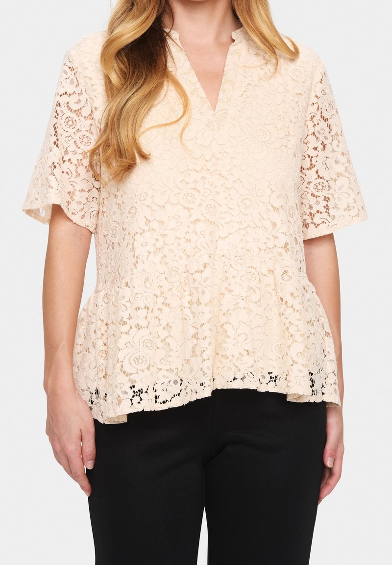 Lace peplum top in light beige with a v-neckline and short sleeves. Features detailed floral patterns and scalloped hem, paired with black trousers.