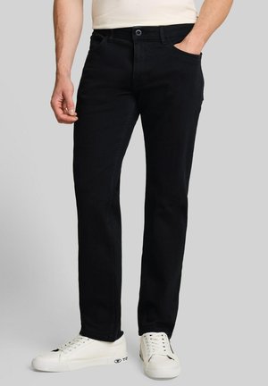 Mann trägt schwarze Jeans mit geradem Bein und weiße Sneaker und steht mit einer Hand in der Tasche vor grauem Hintergrund.