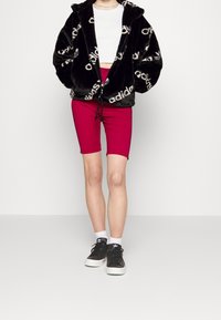 Černá plyšová bunda na zip s bílým logem "adidas", doplněná přiléhavými červenými šortkami a černými teniskami. Jsou viditelné bílé ponožky.