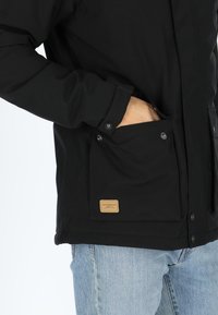 Black jacket with button-up front, side pockets, and a textured fabric. Brown brand label on pocket adds contrast and detail.
