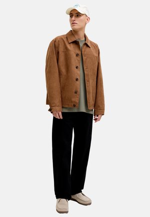 Mann mit beiger Kappe, brauner Jacke mit Knöpfen, grünem Hemd, schwarzer Hose und beigen Schuhen, steht und schaut seitlich vor weißem Hintergrund.