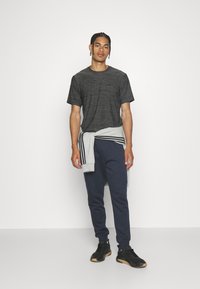 Sort tekstureret sportstrøje, marineblå joggers, grå sweatshirt bundet om taljen, og sorte sneakers med gummisål. Simpel, sporty påklædning.