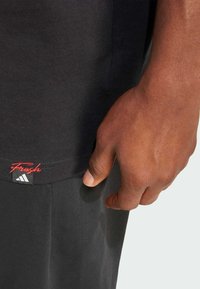 Crni majica od pamuka, s oznakom logoa na rubu s natpisom "Fresh" u crvenoj boji i Adidas emblemom. Tekstura tkanine je glatka.