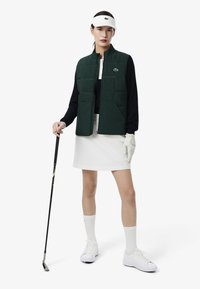 Chaleco acolchado verde oscuro con diseño acolchado, camiseta negra de manga larga, falda blanca, visera blanca y guantes. El modelo sostiene un palo de golf.