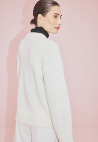 Bela pletena jopica z rebričastim robom in manšetami, z ohlapnim krojem in teksturiranim vzorcem. Ozadje je mehka rožnata barva.