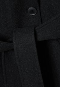 Čierny vlnený kabát s textúrovaným povrchom, s opaskom v páse a okrúhlym zapínaním na gombíky. Tkanina je mäkká a chlpatá.