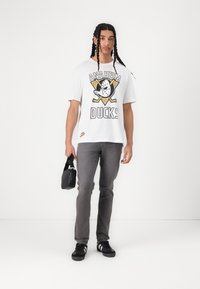 T-shirt graphique blanche avec le design "Anaheim Ducks", jean slim gris, baskets noires et un petit sac noir. Cheveux tressés.
