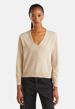 Beige v-hals trui met lange mouwen, zachte textuur en geribde zoom. Gecombineerd met donkere jeans, worden contrasterende kleuren en materialen benadrukt.
