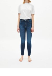 Una persona que lleva una camisa blanca de manga corta y unos jeans ajustados de color azul oscuro, de pie descalzo sobre un fondo blanco.