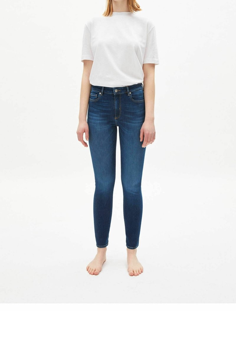 Una persona que lleva una camisa blanca de manga corta y unos jeans ajustados de color azul oscuro, de pie descalzo sobre un fondo blanco.