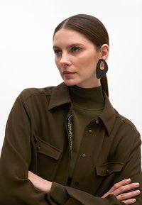 Camicia verde oliva oversize con bottoni e tasche, indossata su un dolcevita verde scuro. Grandi orecchini geometrici neri e dorati.