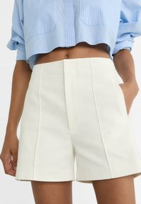 Pantaloni corti plissettati di colore crema con tasche laterali e una texture liscia, abbinati a una camicia corta a righe blu chiaro.