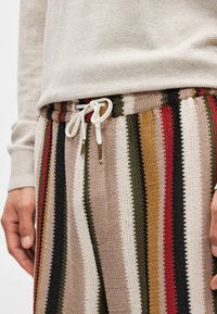 Πολύχρωμοι ριγέ πλεκτοί παντελόνες με κορδόνι στη μέση και χρυσές λεπτομέρειες, που προβάλλουν αποχρώσεις του μπεζ, κόκκινου, πράσινου και μαύρου.