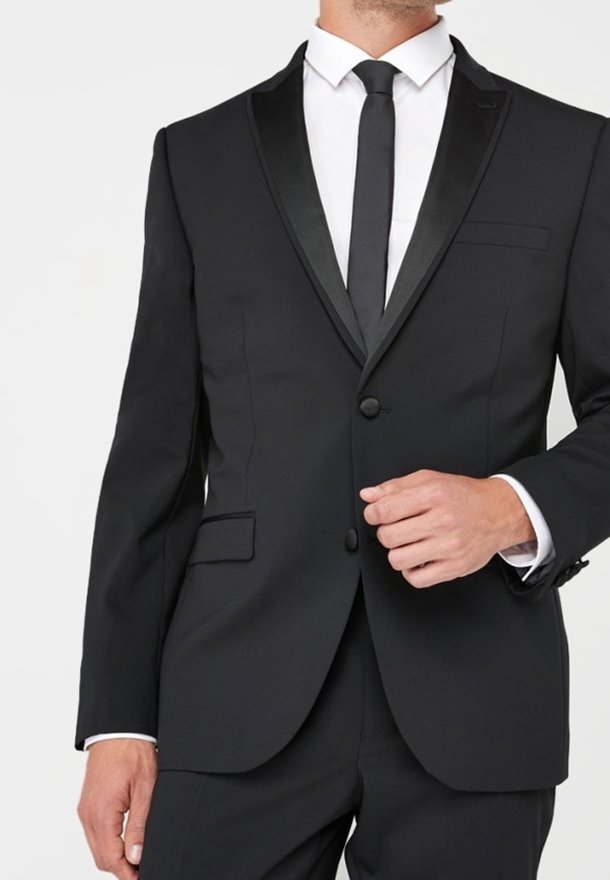 Homme portant un smoking noir avec des revers en satin, une chemise blanche et une cravate noire, une main ajustant le bouton de la veste.