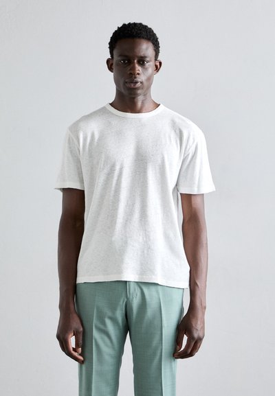 Tricou alb cu maneci scurte și guler rotund, fabricat dintr-un material moale, asociat cu pantaloni personalizați de culoare verde deschis, prezentat pe un fundal neutru.