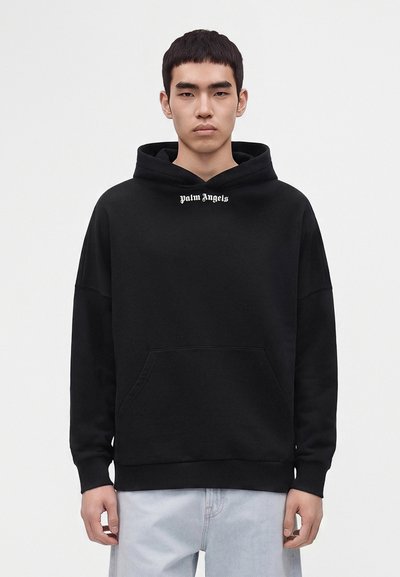 Jeune homme aux cheveux noirs courts portant un sweat à capuche noir Palm Angels et un jean bleu clair sur un fond blanc uni.