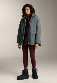 Osoba odjevena u sivu prošivenu jaknu s kapuljačom, smeđi flis prsluk, boje grimiz tajice, sive čarape i crne zimske čizme, stoji na neutralnoj pozadini.