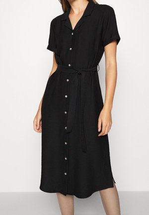 Femme portant une robe midi noire à manches courtes, boutonnée, avec col et ceinture nouée à la taille, debout sur un fond blanc.