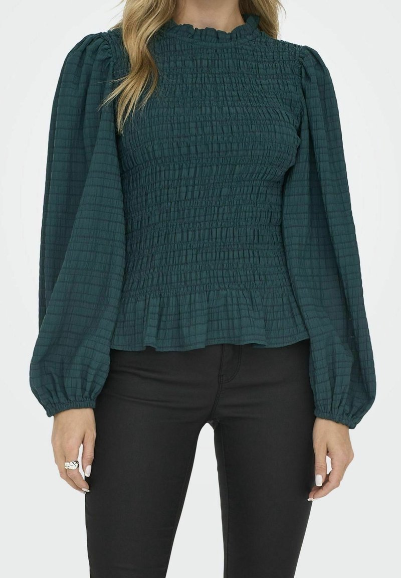 Femme portant un chemisier smocké vert foncé avec des manches longues bouffantes et un pantalon noir ajusté, debout devant un fond uni.