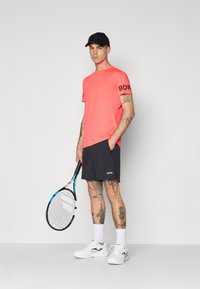Koralinė sportinė marškinė su "BORG" logotipu ant rankovių, juodos šortai su "BORG" logotipu, balti sportbačiai, laikantis teniso raketę.