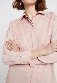 Femme portant un chemisier rose pâle avec des panneaux en dentelle, une boucle d'oreille pendante en perle et une bague en or, bras croisés devant un fond uni.