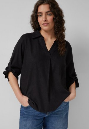 Frau mit lockigem braunem Haar, die eine lockere schwarze V-Ausschnitt-Bluse mit hochgekrempelten Ärmeln und blaue Jeans trägt, steht mit den Händen in den Taschen.