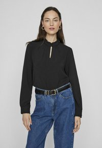Musta pitkähihais blouse, jossa on rypytyskraavu ja avonainen kaula-aukko, yhdistettynä korkeavyötäröisiin sinisiin farkkuihin ja mustaan vyöhön.