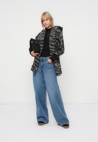 Manteau en fausse fourrure grise avec des rayures noires de tigre, assorti à un haut noir ajusté et un jean bleu à jambes larges. Sac noir tenu dans les bras.