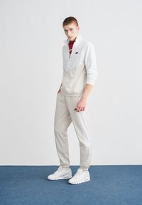 Chaqueta deportiva gris claro de Nike con cuello alto y logo negro, combinada con pantalones deportivos grises a juego y zapatillas blancas, sobre un suelo azul.