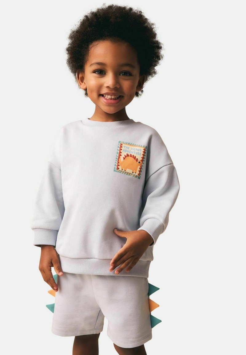Enfant souriant portant un sweat-shirt gris clair avec un écusson de dinosaure et un short assorti avec des pointes de dinosaure colorées sur les côtés.