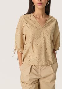 Beige shirt with a V-neck, featuring an eyelet design in a diamond pattern, short puff sleeves with ties, and a textured finish.