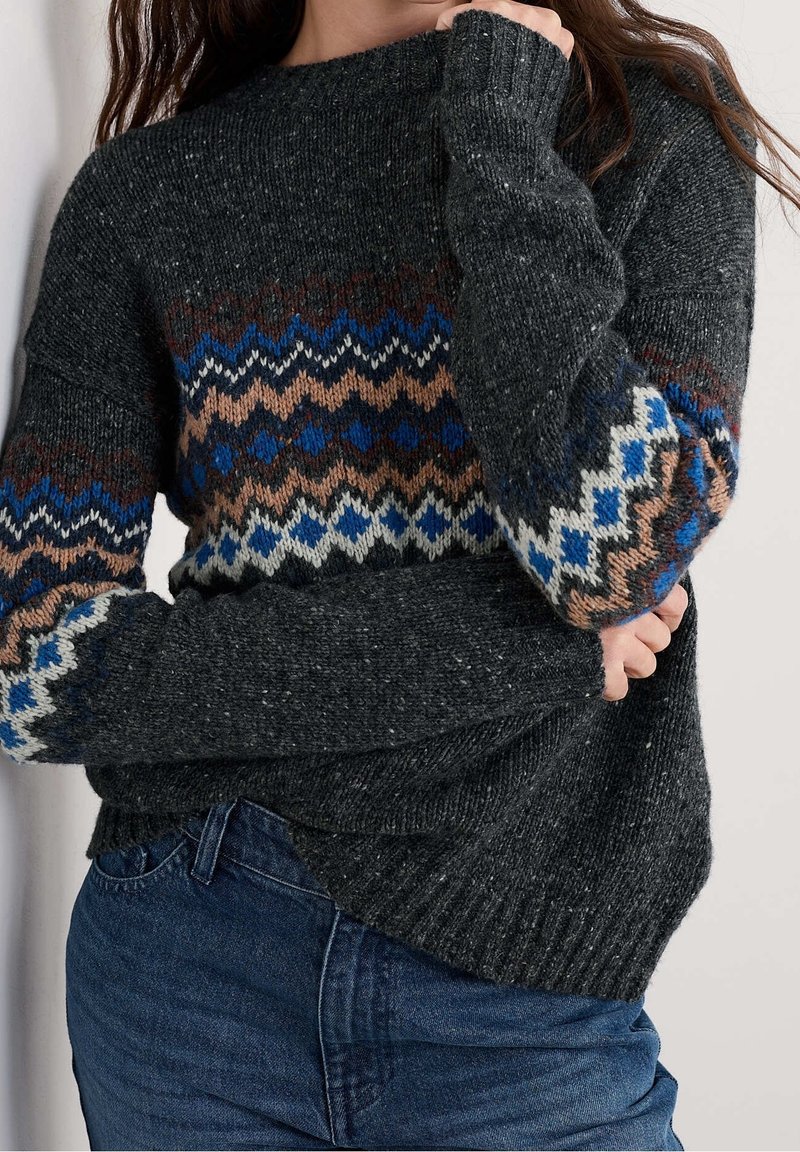 Personne portant un pull en maille gris foncé avec des rayures à motifs bleues, beige, blanches et brunes sur la poitrine et les manches, associé à un jean bleu.