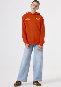 Young woman wearing an orange hoodie with white text, light blue wide-leg jeans, and leopard print sneakers standing against a plain white background.