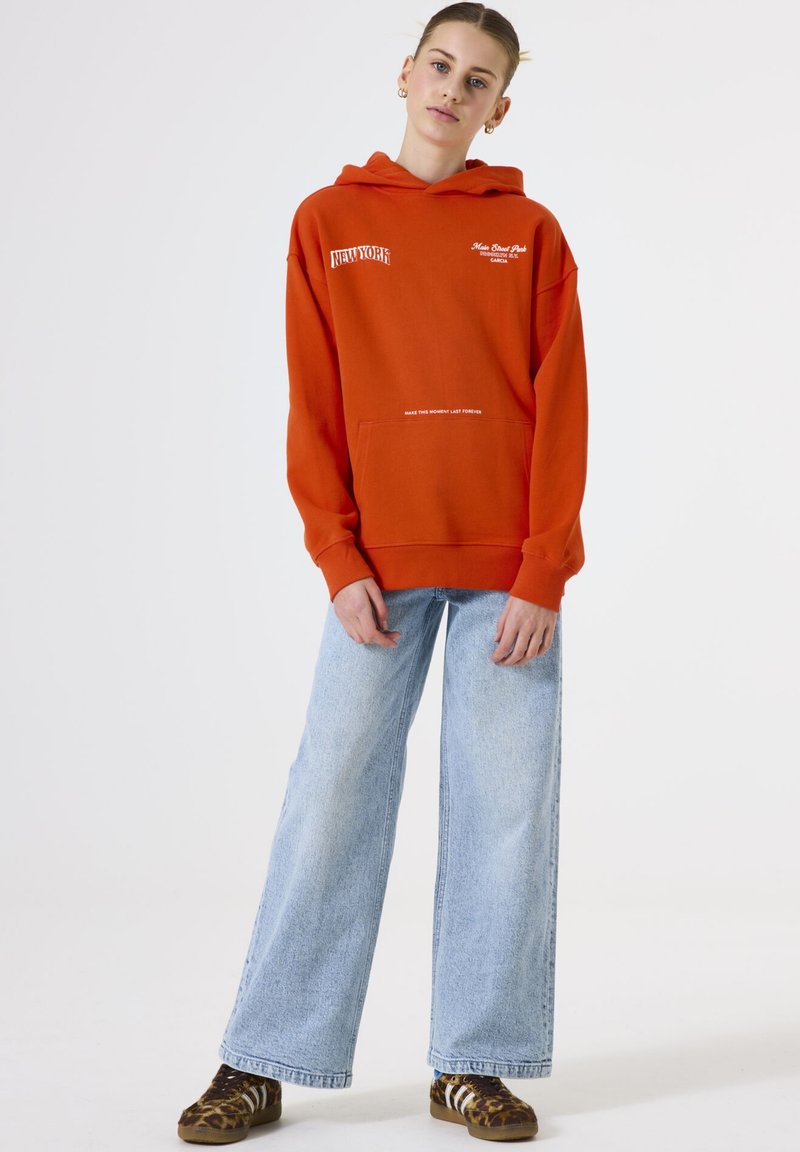 Young woman wearing an orange hoodie with white text, light blue wide-leg jeans, and leopard print sneakers standing against a plain white background.