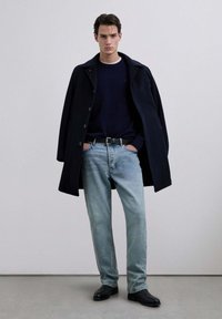 Cappotto di lana blu navy su un maglione blu navy, abbinato a jeans blu chiaro e stivali in pelle nera. Linee pulite e vestibilità rilassata in tutto l'outfit.