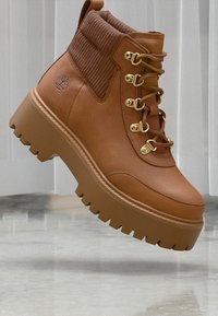 Bottine en cuir brun avec un col en tissu côtelé, des œillets en métal doré et une semelle en caoutchouc épaisse avec des crampons profonds.