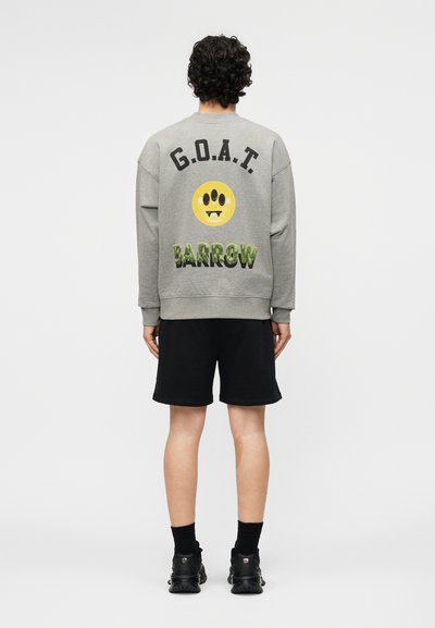 Άτομο στέκεται με την πλάτη φορώντας γκρι φούτερ με το κείμενο "G.O.A.T.", κίτρινο χαμογελαστό πρόσωπο με τρία μάτια και πράσινο κείμενο "Barrow", συνδυασμένο με μαύρο σορτς.