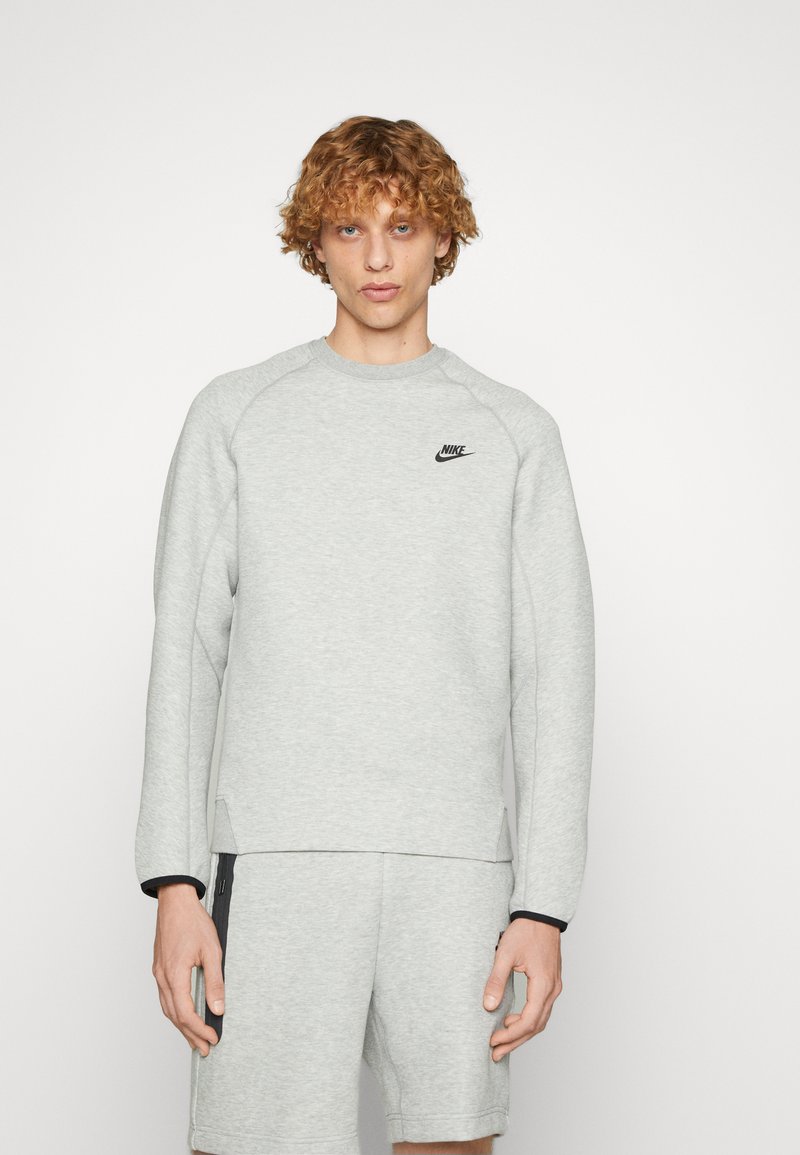 Sweatshirt gris en tissu doux avec des manches longues, un col côtelé et des fentes latérales. Il arbore un petit logo Nike noir sur la poitrine.