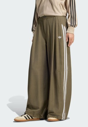 Person iført brunternede bukser med vide ben, hvide sidelister og Adidas-logo, kombineret med beige Adidas-sko med sorte striber.