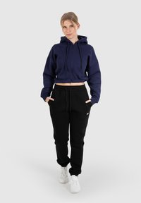 Felpa blu navy con cappuccio, zip frontale e cordini, abbinata a pantaloni della tuta neri con tasche. Il modello indossa sneakers bianche.