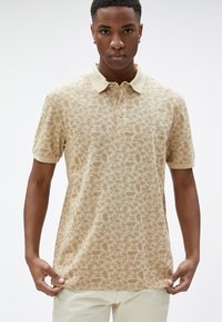 Camisa polo beige con estampado floral, de manga corta y cuello marrón claro. Material de algodón con una textura suave. Tapeta de dos botones.