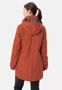 Giacca impermeabile color ruggine con cappuccio, polsini elasticizzati e vita regolabile con cordino. Il materiale sembra liscio con una fodera interna texturizzata.