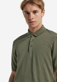 Olivgrön polo med strukturerad tyg, med krage, tre knappar och korta ärmar.