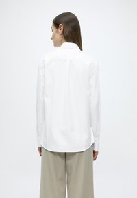 Λευκ ό βαμβακερό πουκάμισο με μακριά μανίκια, κλασικό γιακά, πτυχώσεις στο πίσω μέρος και καμπύλη γραμμή στο κάτω μέρος, συνδυασμένο με ανοιχτό μπεζ παντελόνι φαρδιάς γραμμής.