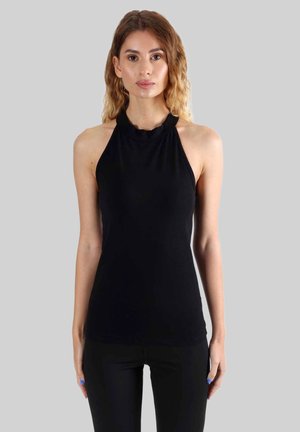 Jeune femme aux cheveux châtain clair portant un haut noir sans manches à col halter et un pantalon noir, debout devant un fond gris uni.