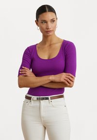 Top ajusté mauve à côtes avec un décolleté plongeant et des manches courtes, associé à un jean clair et une ceinture brune avec des éléments en métal argenté.