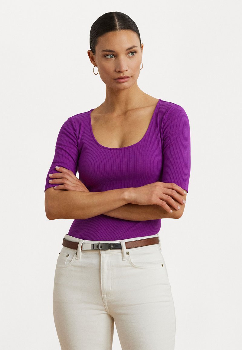 Top ajusté mauve à côtes avec un décolleté plongeant et des manches courtes, associé à un jean clair et une ceinture brune avec des éléments en métal argenté.