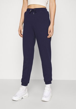Person iført navyblå joggers med snøre, hvid crop top, hvide sokker og hvide træningssko, der står på en grå gulv.