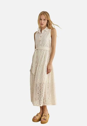 Mujer con vestido midi blanco sin mangas con cuello, encaje calado, botones y cinturón en la cintura, combinado con sandalias de plataforma color tostado.